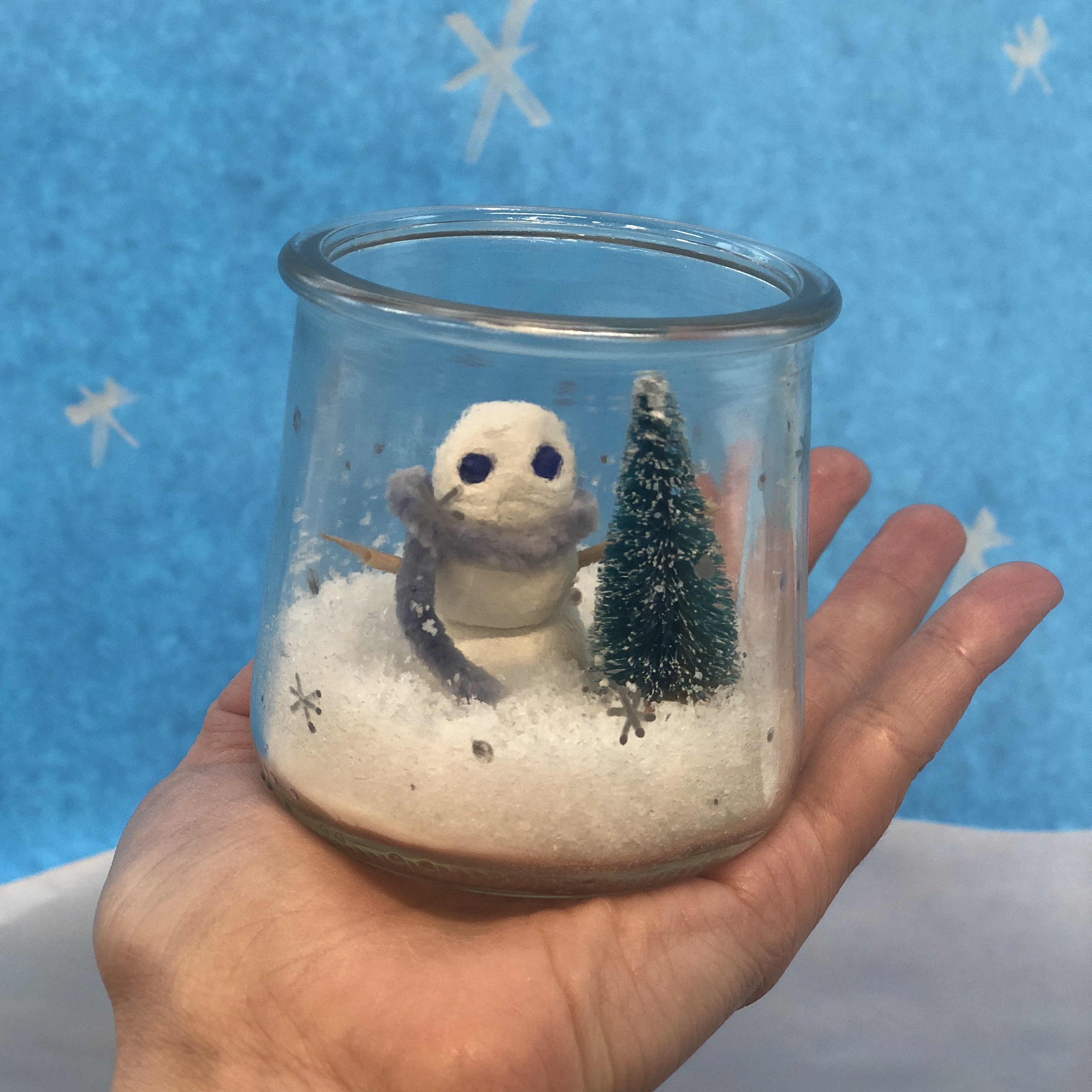 Photo of a snow scene with a small snowman wearing a scarf, fake snow, and a tiny tree, inside of a small jar.