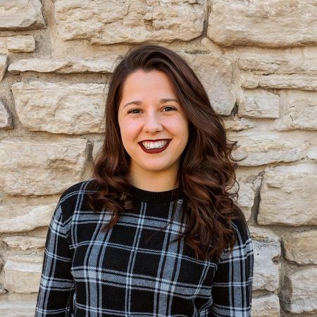 Anna Sortino Author Photo - woman with brown curly hair and wearing a blue plaid sweater standing in front of a stone wall. 